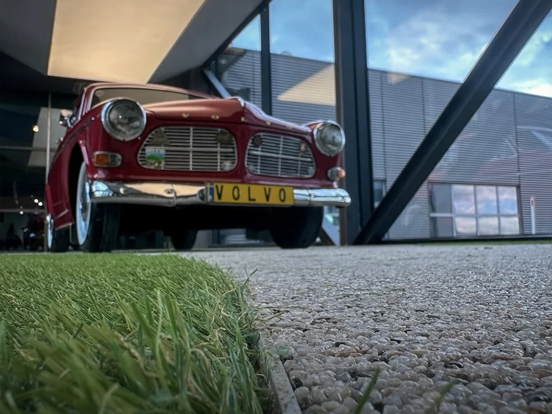 Voiture ancienne rouge garée sur un sol de pierre en granulats, au ras d’une bande de gazon, dans un hall vitré