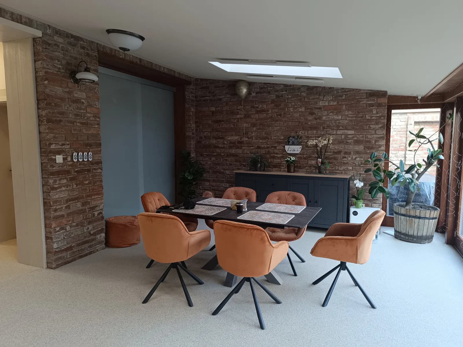 Salle à manger au plancher continu de pierre claire, mur de brique, table et chaises orange devant une baie vitrée